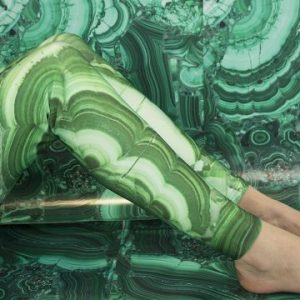 A still image from Emily Speed's video work Rooms Designed for a Woman, showing legs, sat on the floor, with bent knees and bare feet. The legs are clad in green tight fitted leggings with a geological print, which matches the backdrop filling the background of the images.