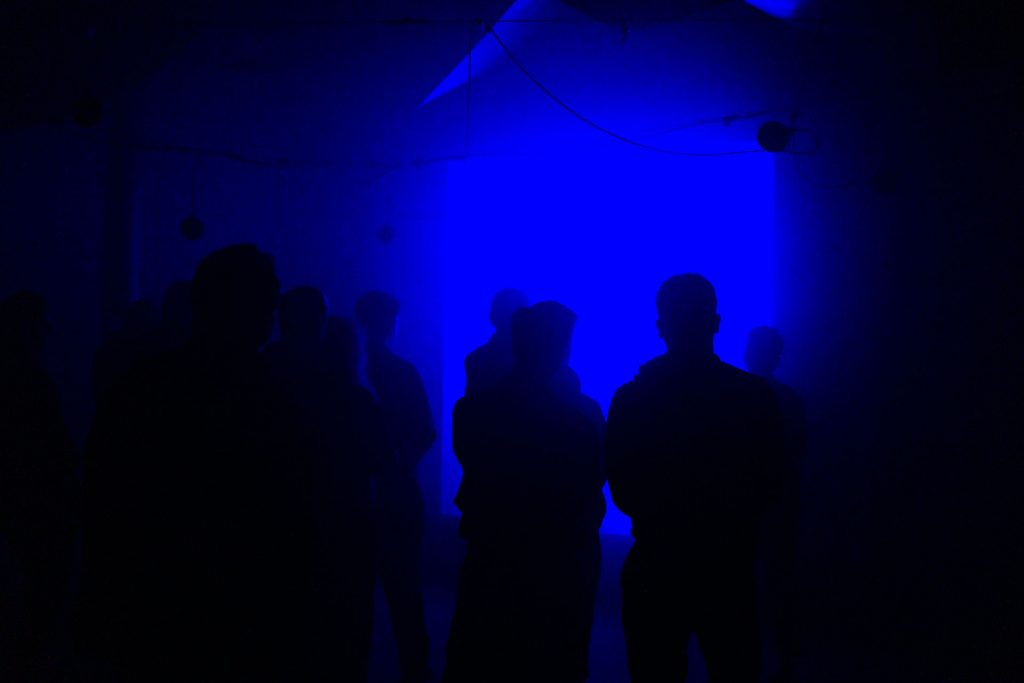 A photograph showing dark sillouttes, backlit in blue, as the figures watch The Conductor by Mishka Henner, in the reverberation chamber at the University of Salford.