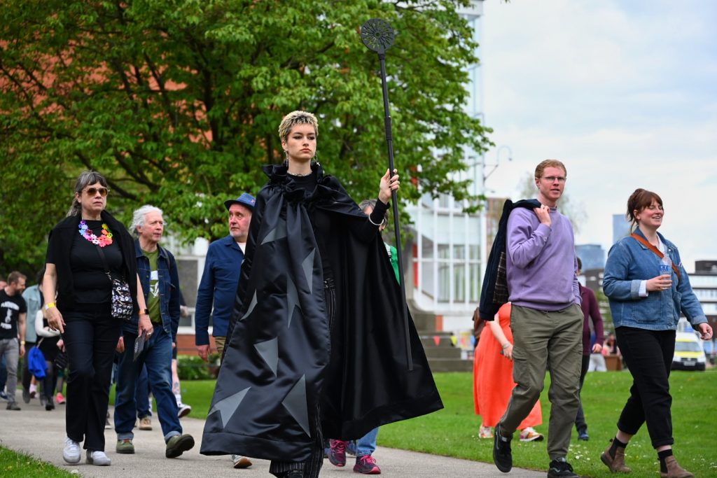 The Protector, dressed in a dark cloak and carying a long staff leads the audience across the University of Salford Campus.