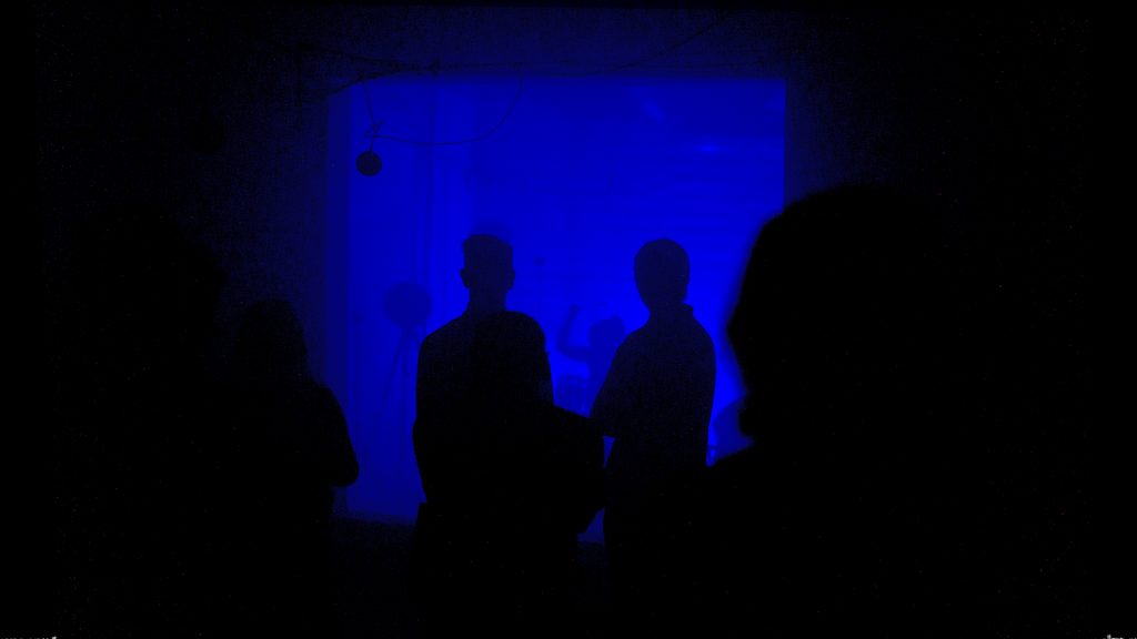 A photograph showing dark sillouttes, backlit in blue, as the figures watch The Conductor by Mishka Henner, in the reverberation chamber at the University of Salford.