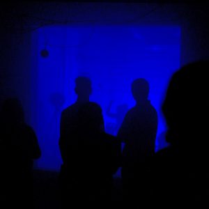 A photograph showing dark sillouttes, backlit in blue, as the figures watch The Conductor by Mishka Henner, in the reverberation chamber at the University of Salford.
