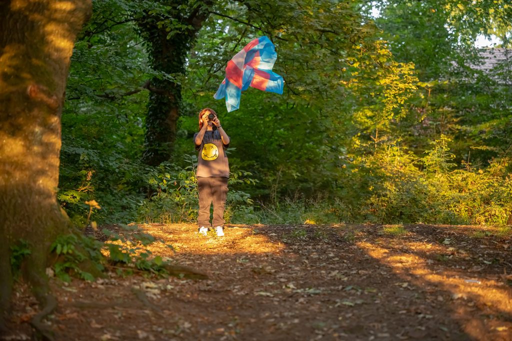 A photograph showing a young person in nature, using a DSLR camera to take a picutre of a transgender pride flag, floating in the air. 