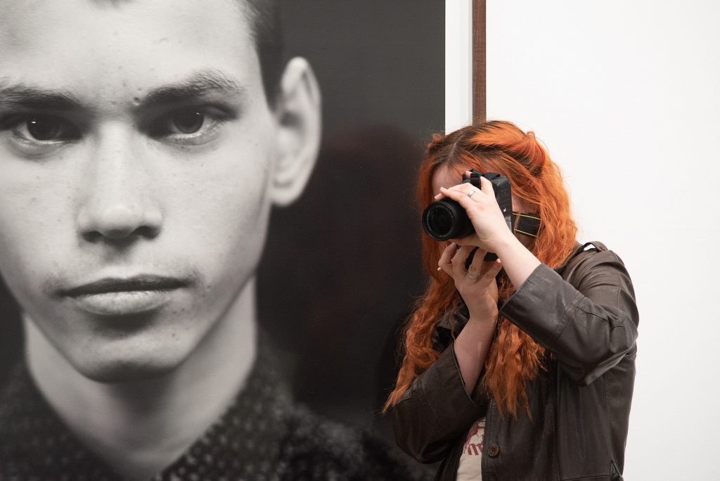 An Our Time, Our Place paricpant with long red hair, holds a DSLR camera up to their eye, while standing infront of a large scale black and white portrait of a young man from the Bank Top series.