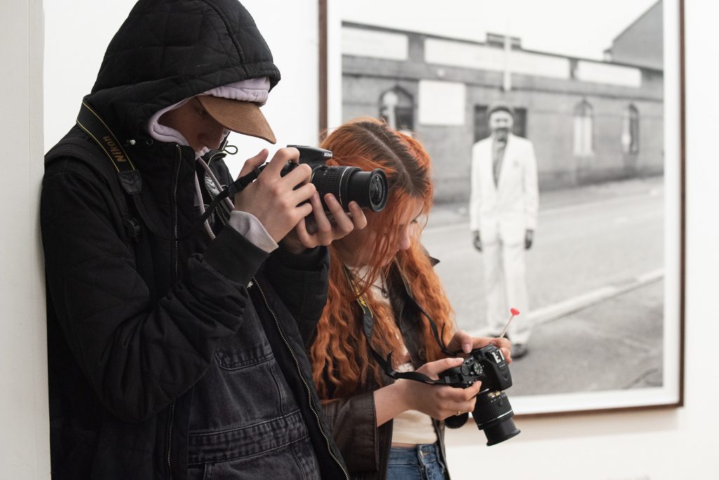 Two young people with cameras, looking at their work and taking images in front of Craig Easton's large, black and white Photographs. 
