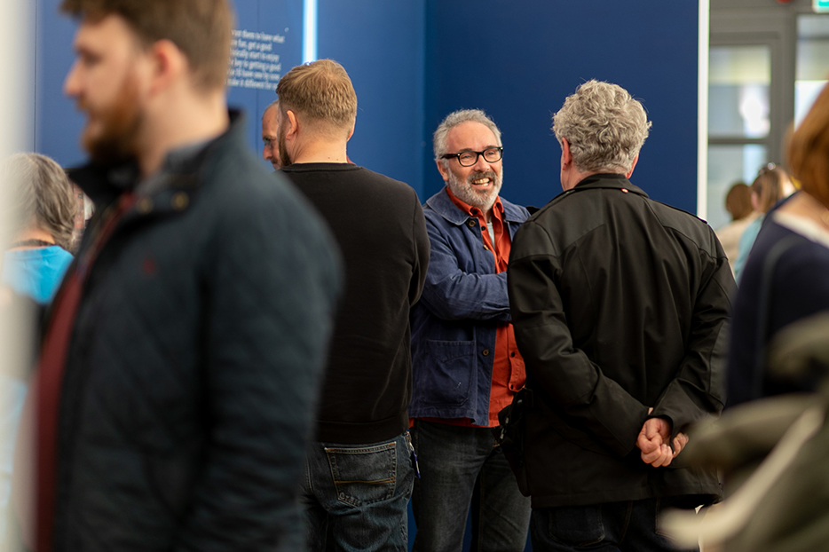 Craig Easton discussing his work with visitors to his exhibition Is Anybosy Listening? In Blackpool. He stands against a navy blue wall.