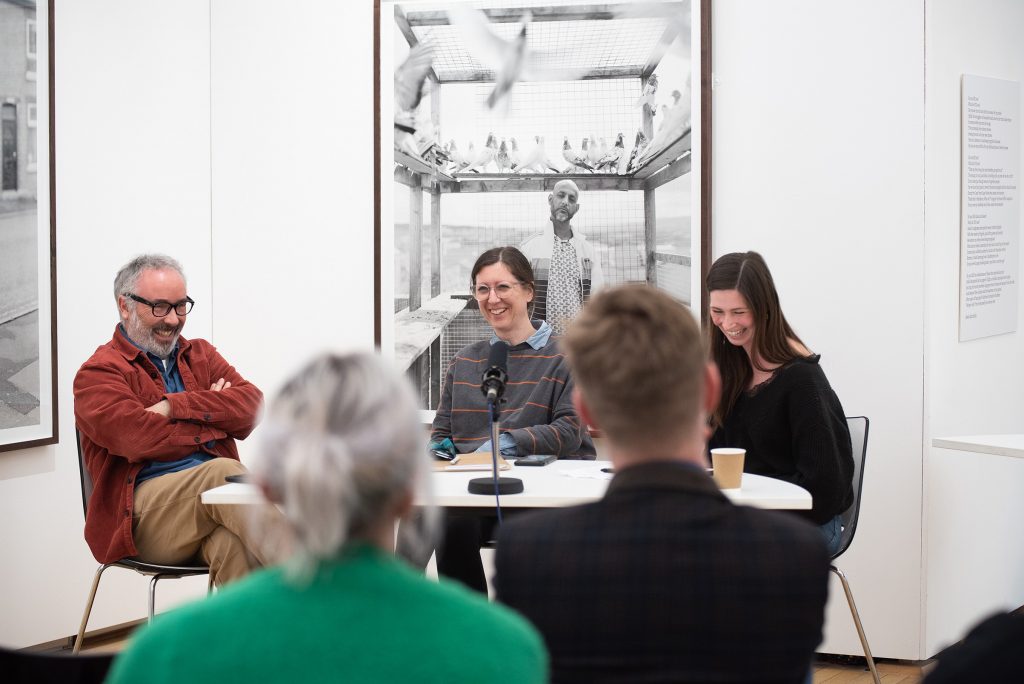 Craig Easton, Laura Jamieson and Liz Wewiora laughing together while sat around a table in front of Easton's large black and white photographs installed at Blackpool School of Art.