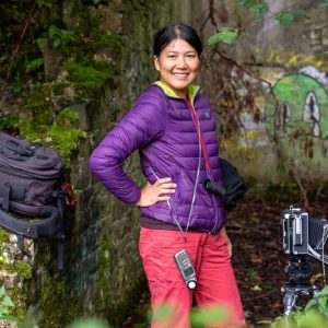 an image of artist yan wang preston. She stands in a green natural space, facing the camera. She wears purple and pink outdoor clothing and has pieces of audio visual recording equipment.