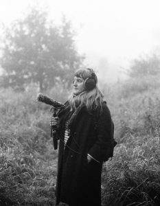 a black and white image of a woman stood infront of trees, she wears headphones and holds a microphone for sound recording. The subject is Hayley Suviste, a sound artist.