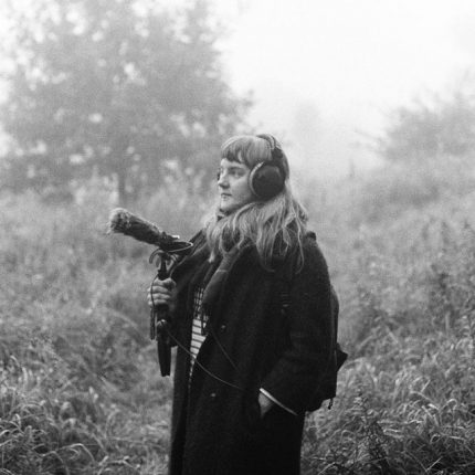 a black and white image of a woman stood infront of trees, she wears headphones and holds a microphone for sound recording. The subject is Hayley Suviste, a sound artist.