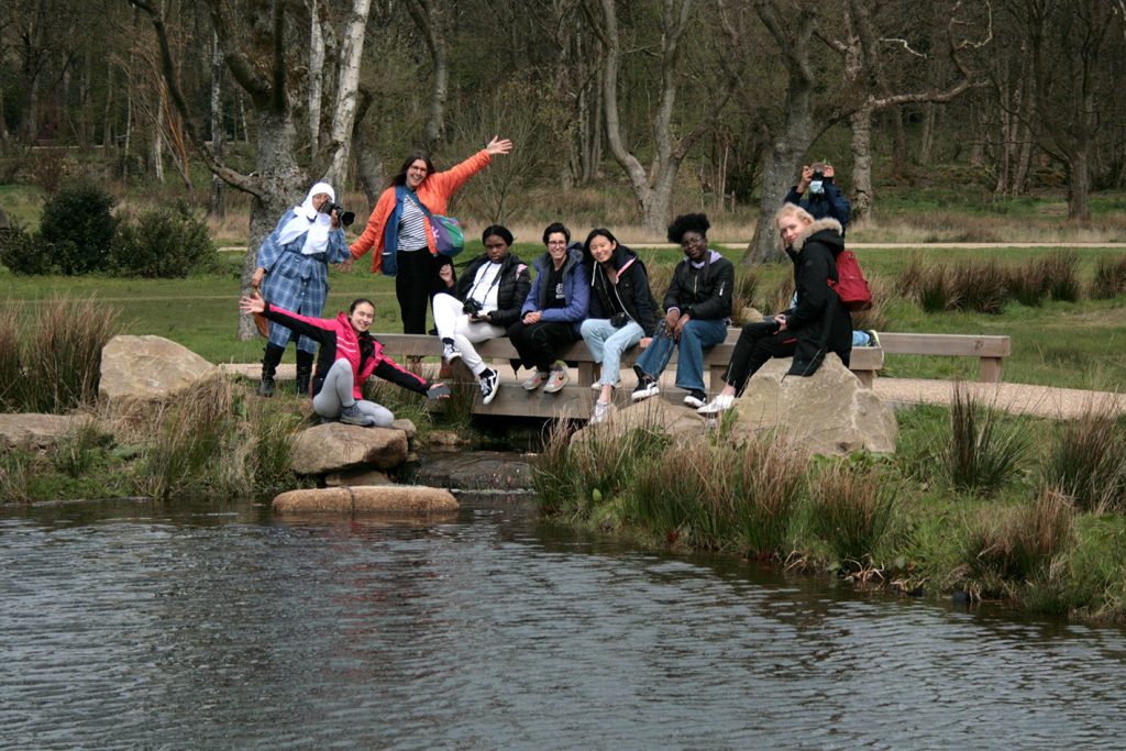 a group of young people sat by a lake in natural surroundings at RHS Garden Bridgewater, they pose for the camera and celebrate