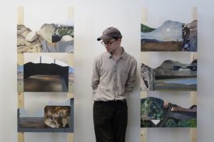 an artist stands between two of his mixed media artworks, against a white wall.