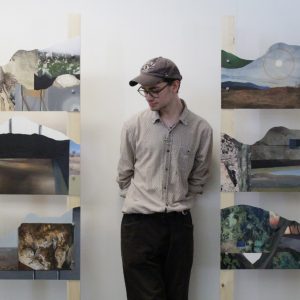 an artist stands between two of his mixed media artworks, against a white wall.