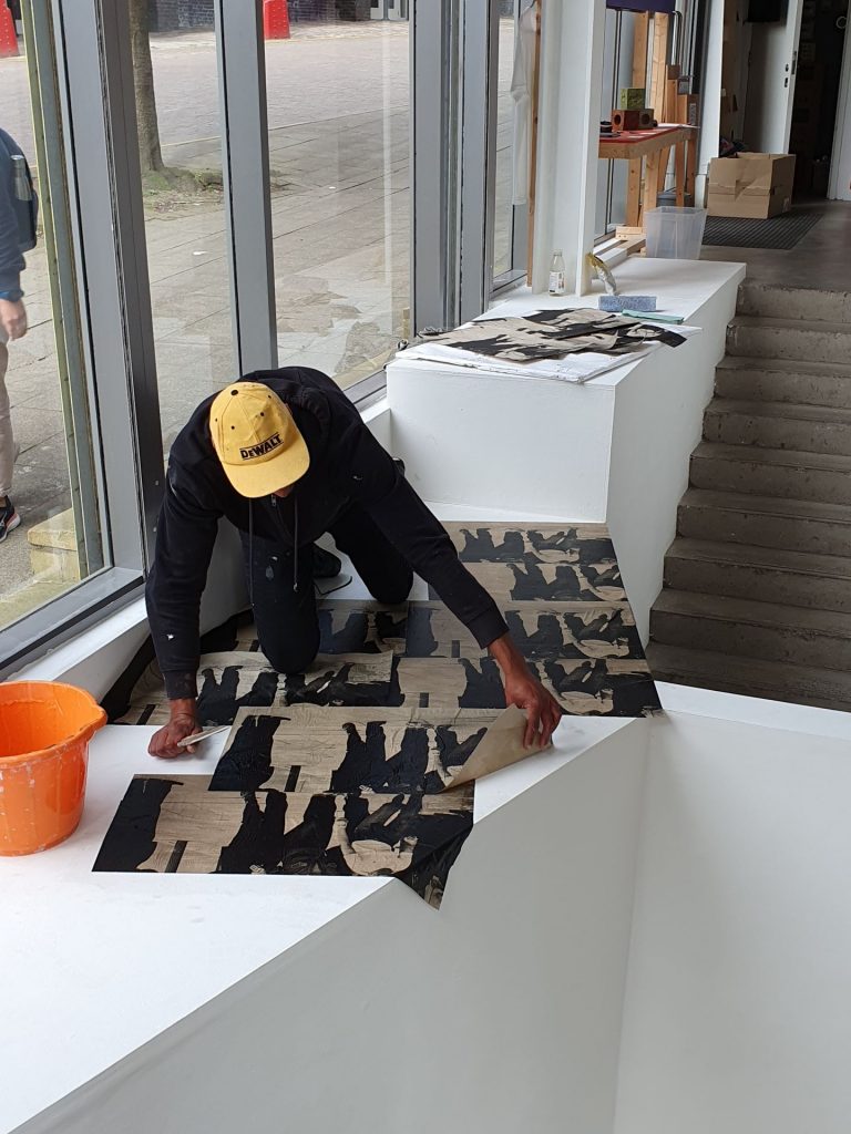 Artist Theo Simpson in a Yellow DeWalt branded baseball cap installing his wallpaper works on the floor of Castlefield Gallery.