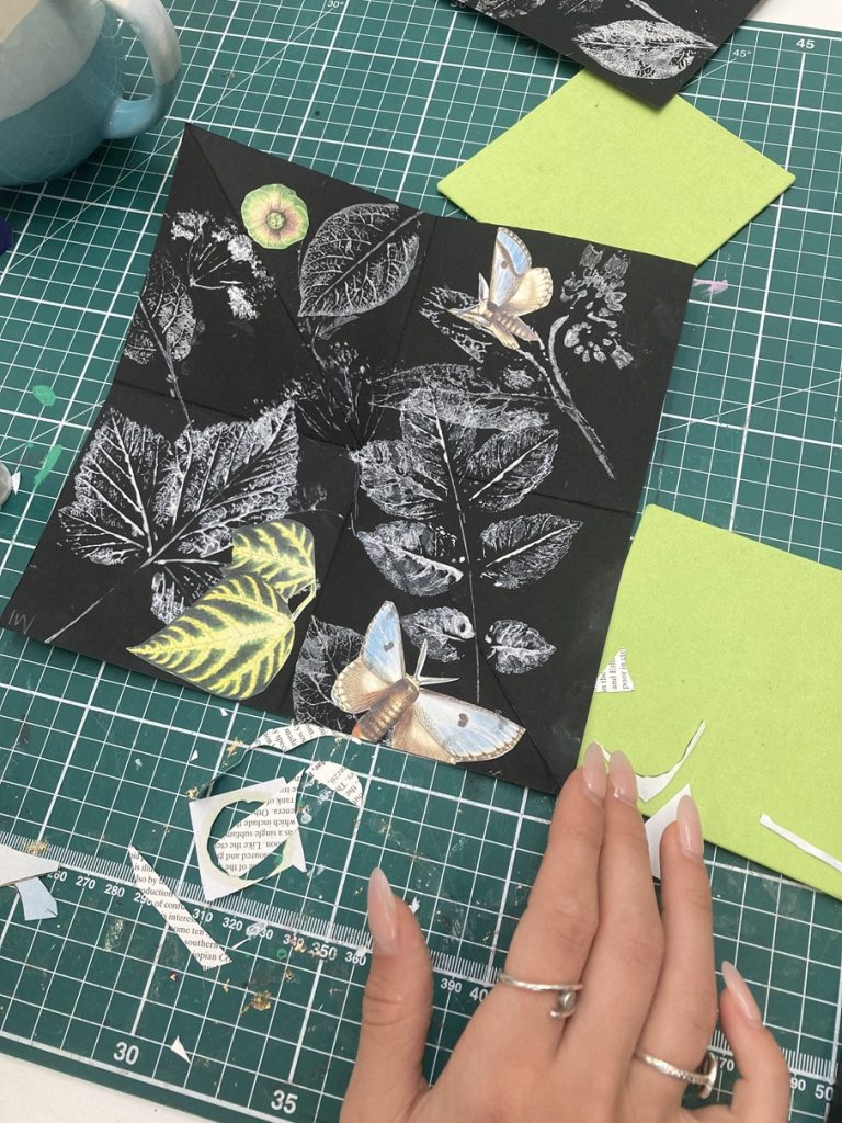 A tabletop with a cutting board, and pieces of coloured paper. A paper collage is in progress, with images of leaves.