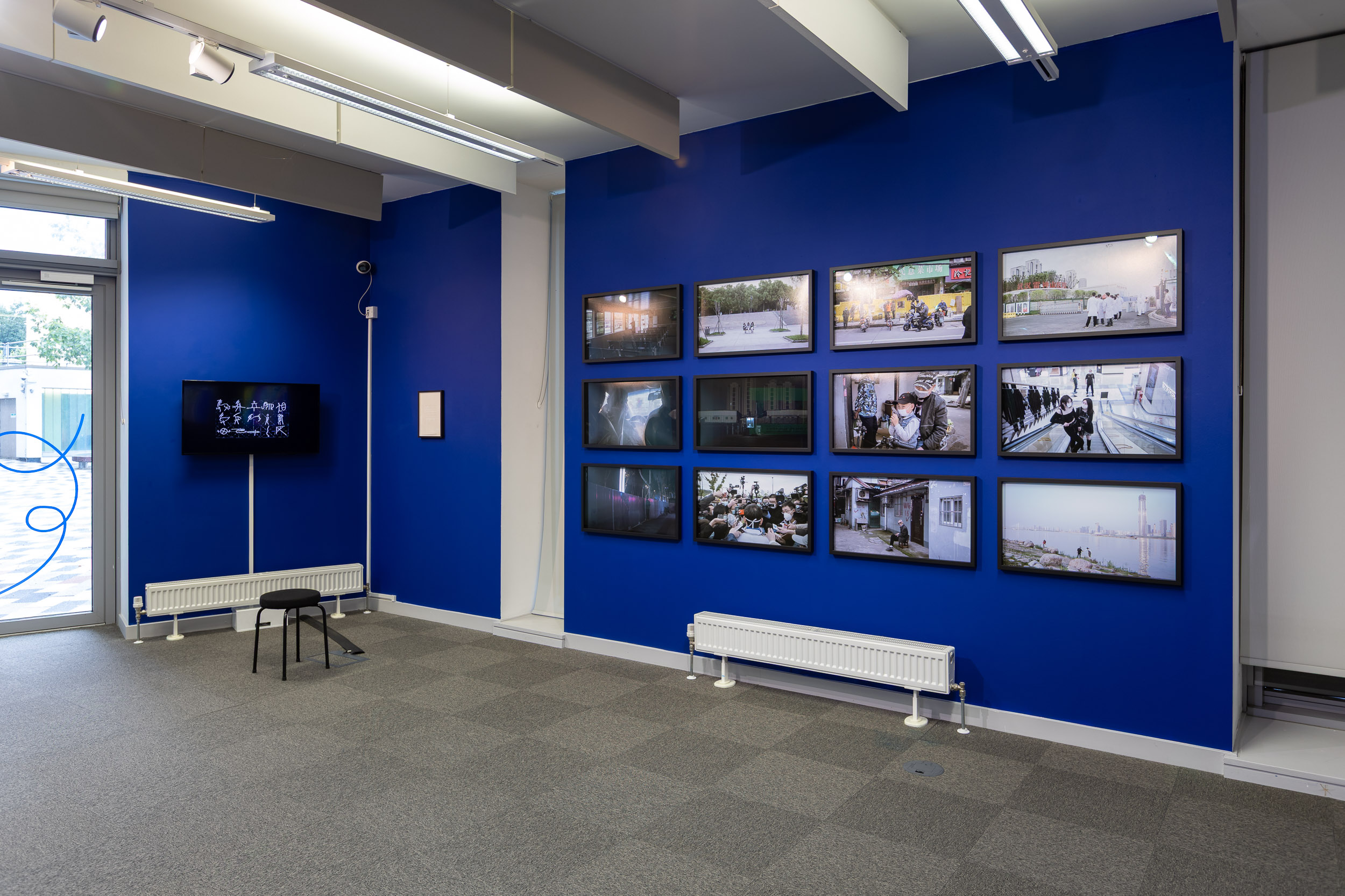 An install photograph of the 'Mediated Realities' exhibition in the New Adelphi Exhibition Gallery. The walls are painted a deep electric blue. From left to right there is: Artwork - Sun Xun's "A War About Chinese Words" (2005). A hand-drawn animation presented on a 42inch TV screen. The animation has a black background with simple white drawn images over the top. It gives the impression of chalk on a very black chalkboard. Artwork - Thomson & Craighead's "The War On Terror" (2016). This work is a small text work in a black frame. The text was written using a typewriter and shows the words "war hero torrent" change into "the war on terror" over 7 lines. The text is typed in black ink, however "the war on terror" line is in red. Artwork - Wu Yue's "a transparent separation" (2020). These are framed 35mm anaolgue photographs taken in Wuhan during the Pandemic in 2020. There are 12 of these photographs in total filling the left wall in a grid-like-fashion.