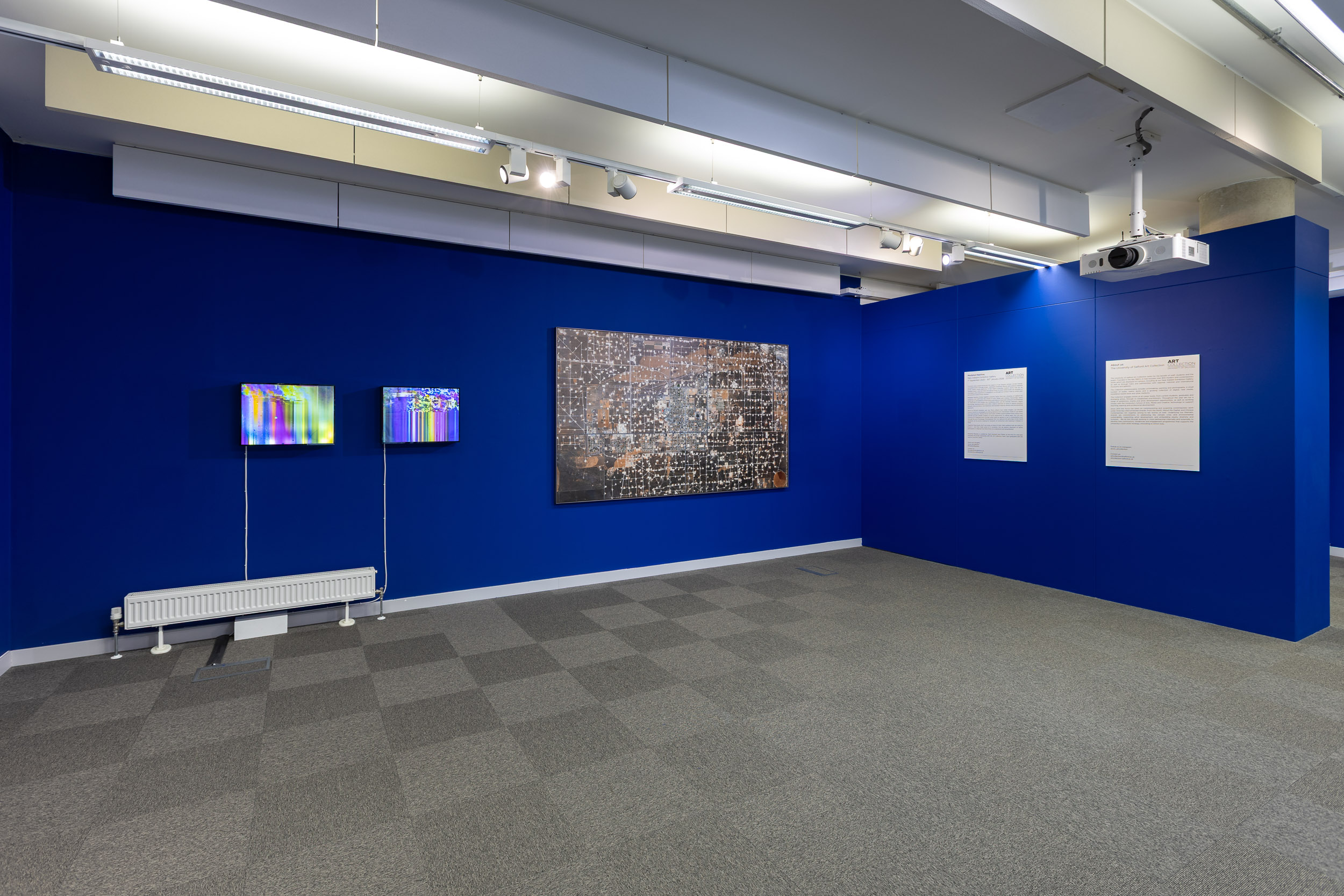 An install photograph of the 'Mediated Realities' exhibition in the New Adelphi Exhibition Gallery. The walls are painted a deep electric blue. From left to right there is: Artwork - Thomson & Craighead's "Corruption" (2014). There are 2 of these lightboxes on the wall with a white cable coming down from the bottom left of each in a straight line. The lightboxes themselves show a glitched, corrupted video files with an eclectic mix of yellows, pinks, blues, and greens. Artwork - Mishka Henner's "Wasson Oil and Gas Field, Yoakum County, Texas" (2013-14). This is a huge photograph on the wall showing hundreds of stitched together satellite images. The images show the effect that the oil industry has had on the surrounding area - leading to the ground being all different shades of brown and muddy greens - the only white sections are where the oil drills are located - and there are hundreds of them filling the image. Then there are 2 A1 information panels on a separate wall detailing the exhibition, and some information about the University of Salford Art Collection.
