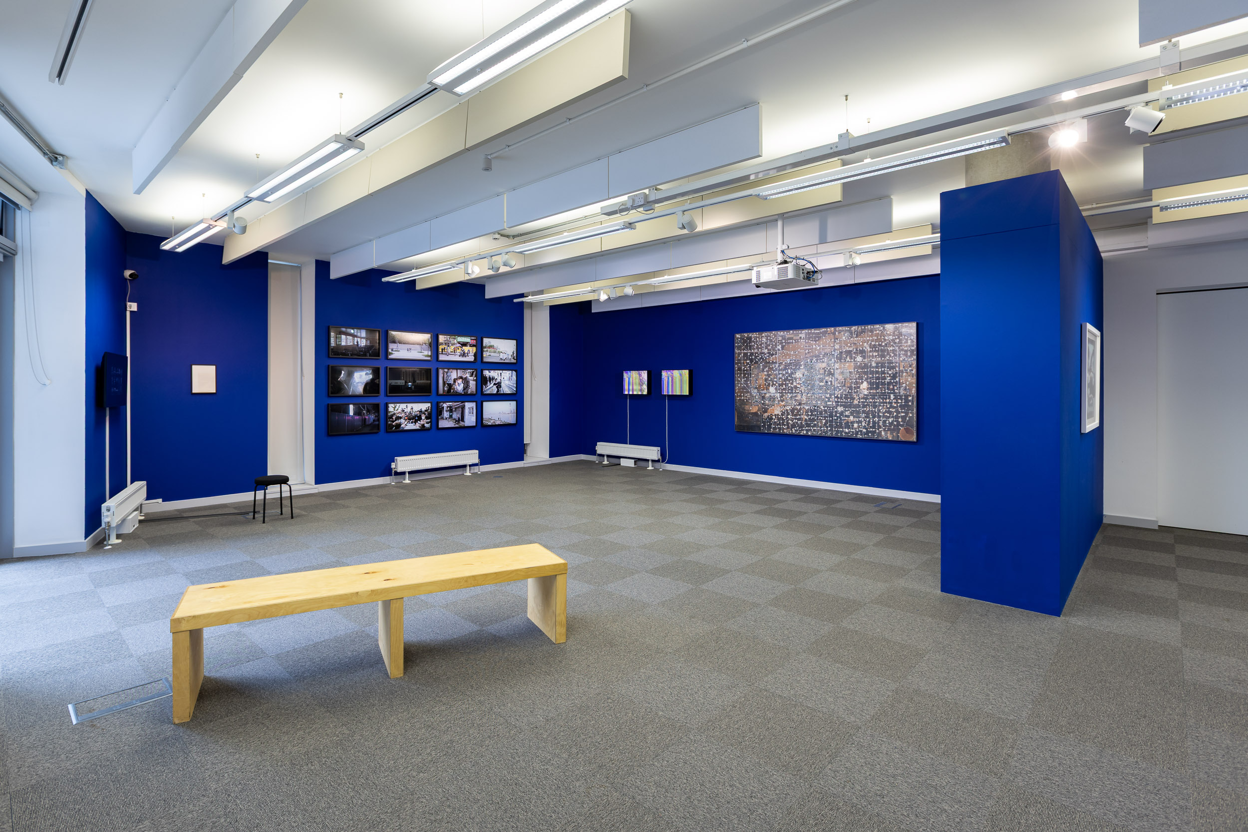 An install photograph of the 'Mediated Realities' exhibition in the New Adelphi Exhibition Gallery. The walls are painted a deep electric blue. From left to right there is: Artwork - Sun Xun's "A War About Chinese Words" (2005). A hand-drawn animation presented on a 42inch TV screen. The animation has a black background with simple white drawn images over the top. It gives the impression of chalk on a very black chalkboard. Artwork - Thomson & Craighead's "The War On Terror" (2016). This work is a small text work in a black frame. The text was written using a typewriter and shows the words "war hero torrent" change into "the war on terror" over 7 lines. The text is typed in black ink, however "the war on terror" line is in red. Artwork - Wu Yue's "a transparent separation" (2020). These are framed 35mm anaolgue photographs taken in Wuhan during the Pandemic in 2020. There are 12 of these photographs in total filling the left wall in a grid-like-fashion. Artwork - Thomson & Craighead's "Corruption" (2014). There are 2 of these lightboxes on the wall with a white cable coming down from the bottom left of each in a straight line. The lightboxes themselves show a glitched, corrupted video files with an eclectic mix of yellows, pinks, blues, and greens. Artwork - Mishka Henner's "Wasson Oil and Gas Field, Yoakum County, Texas" (2013-14). This is a huge photograph on the wall showing hundreds of stitched together satellite images. The images show the effect that the oil industry has had on the surrounding area - leading to the ground being all different shades of brown and muddy greens - the only white sections are where the oil drills are located - and there are hundreds of them filling the image. Artwork - Richard Hamilton's "Kent State" (1970). It is a screenprint framed in white. The image in this print was taken from televised news coverage of the Kent State University tragedy, broadcast on television by the BBC. The dominant colour is the black of the shadows and edges of the TV screen, with highlights of white on the ground and the body, de-saturated blues as a mid-tone, and red depicting the bullet wound. This is a wide shot image with a bench in the foreground.