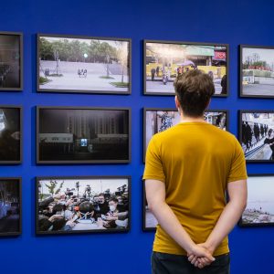 Photograph of Team Assistant Sam Parker, shot from behind, looking at the grid of Yue Wu's photographs as part of the 'Mediated Realities' Exhibition.