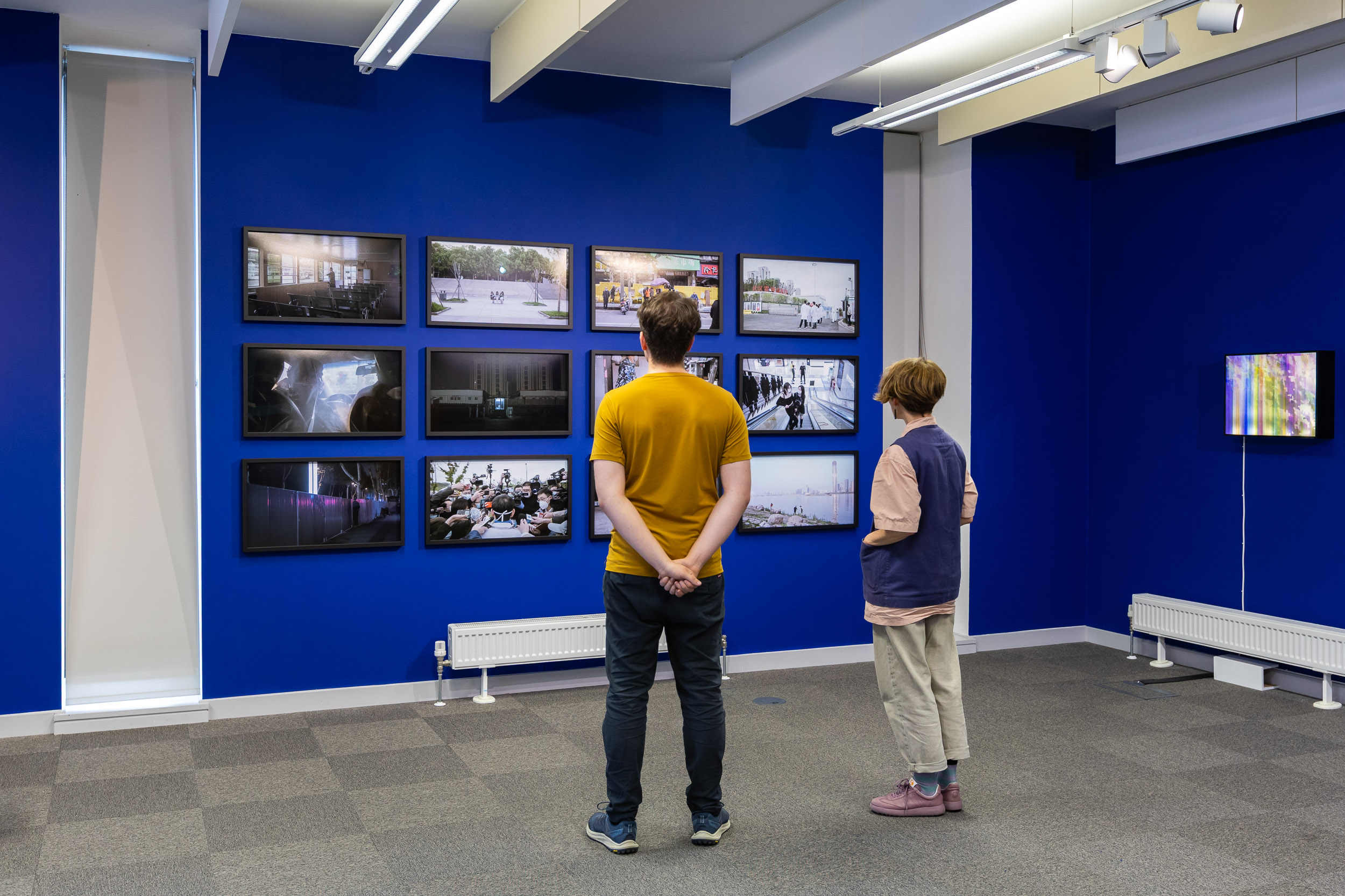 An install photograph of the 'Mediated Realities' exhibition in the New Adelphi Exhibition Gallery. 2 people are photographed looking at Wu Yue's artwork; UoS AC Team Assistant and Curator of this exhibition; Sam Parker, wearing black pants, blue shoes, and a mustard yellow top - fluffy hair as per usual. Alongside him is artist Chrystal Cherniwchan, wearing beige pants, de-saturated purple shoes, and a blue waistcoat over beige t-shirt. The walls are painted a deep electric blue. Artwork - Wu Yue's "a transparent separation" (2020). These are framed 35mm anaolgue photographs taken in Wuhan during the Pandemic in 2020. There are 12 of these photographs in total filling the left wall in a grid-like-fashion.