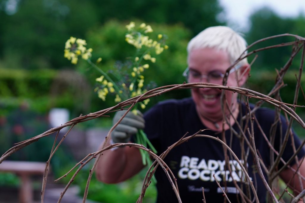 A member of the community looking very happy and exciting to be holding a bunch of yellow flowers, wearing gardening gloves, willow plant in the foreground.