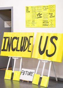an image showing bright yellow placards leaning against a white wall. The text in large black capital letters reads "include us"