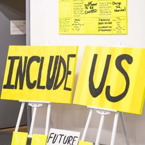 an image showing bright yellow placards leaning against a white wall. The text in large black capital letters reads "include us"