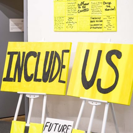 an image showing bright yellow placards leaning against a white wall. The text in large black capital letters reads "include us"