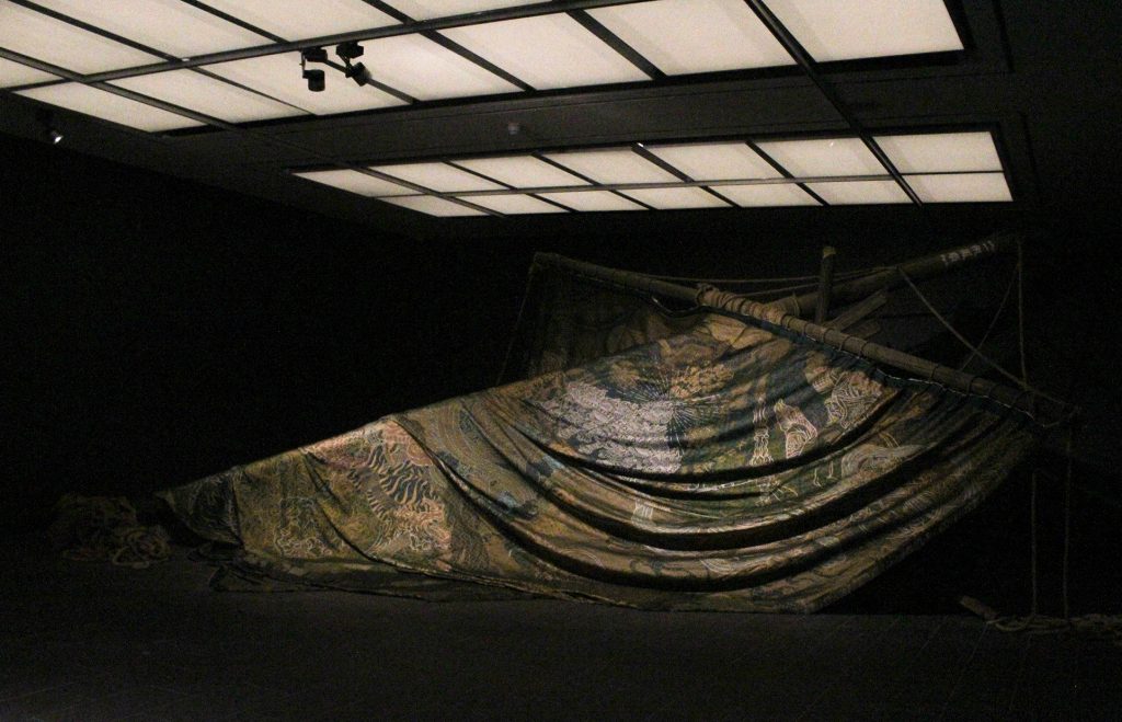 a fallen ship's mast in a darkened gallery space, the sail is a richly embroidered tapestry.