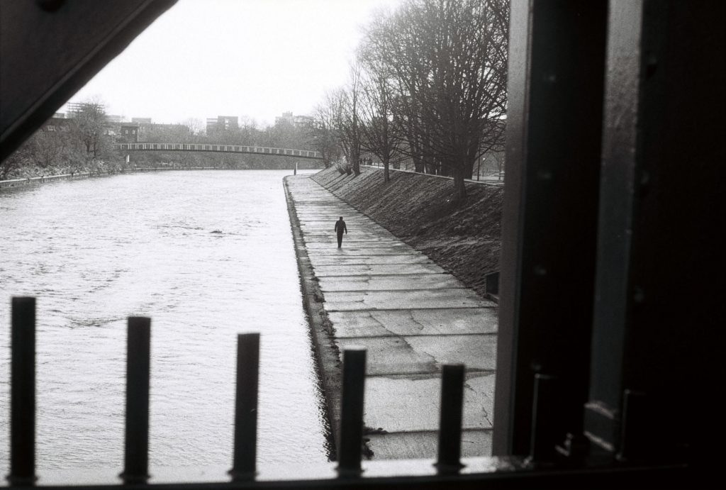 Black and white image taken from behind a gate, looking out onto a river with a walkway running alongside.