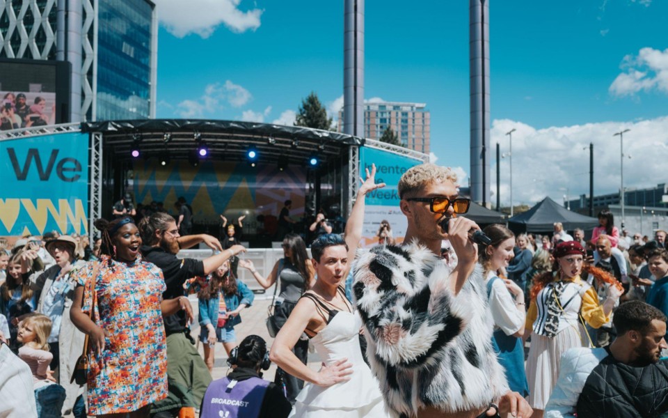Person with a microphone, a large crowd of people and stage behind them on a sunny day.