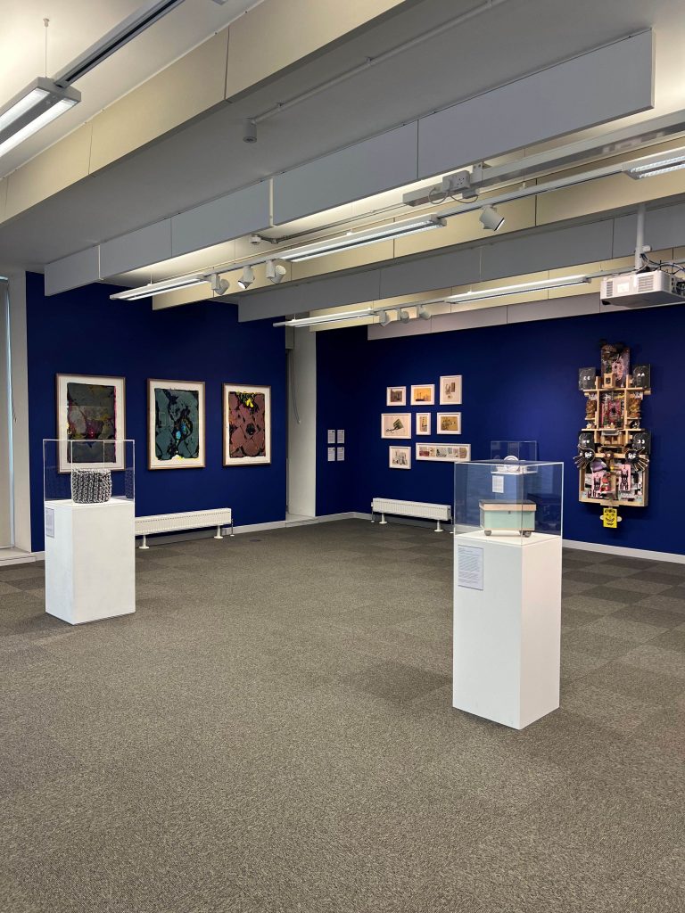 17 different artworks/artwork series' against a blue gallery wall and on top of plinths.