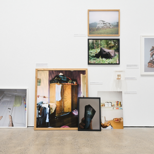 Photographs and archival photographs of families and areas are displayed on the wall and leaning on the floor of a gallery space. Images are from various eras, some in colour and some in black and white.