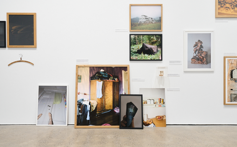 Photographs and archival photographs of families and areas are displayed on the wall and leaning on the floor of a gallery space. Images are from various eras, some in colour and some in black and white.