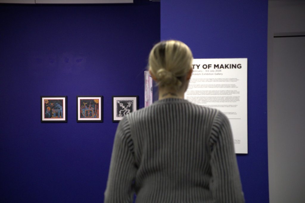 A person views an information panel in a blue painted gallery space, the information title is City of Making, three small collaged artworks are to the left of the person.