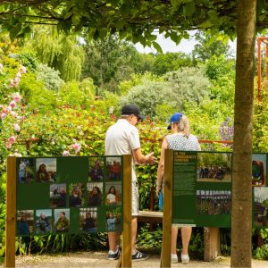 Two people standing the middle of a garden, with bushes and flowers, there is picture boards in front of them.